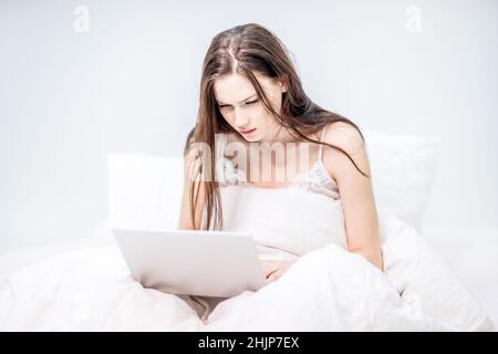 Guten Morgen. Die Frau arbeitet zu Hause. Ein brünette Mädchen sitzt in ihrem Schlafanzug auf einem weißen Bett und hält einen Laptop in den Händen. Entfernt. Stockfoto