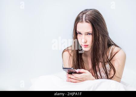 Tag frei. Lesen Sie die Neuigkeiten. Legen Sie sich auf das Bett. Guten Morgen. Eine hübsche Brünette sitzt im Bett, bedeckt mit einer Decke und hält ein Telefon in den Händen. Stockfoto
