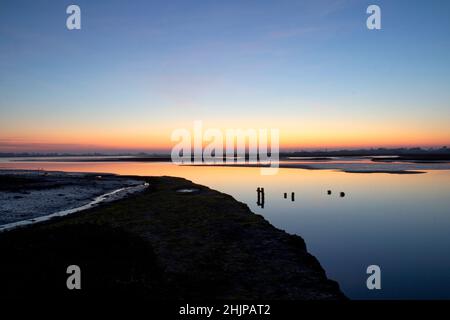 Sonnenuntergang im Naturschutzgebiet Pagham Harbour West sussex bei Ebbe mit Grasnadelgebiet auf der linken Seite und alten Pfosten, die aus dem Wasser ragen. Stockfoto