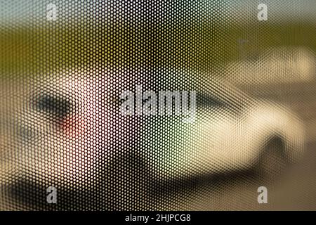 Maschenstruktur auf Glas des Busses. Oberflächendetails von kleinen Löchern. Verschwommener Hintergrund, der durch kleine Löcher geschossen wird. Texturierter Film auf Glas gestreckt. Stockfoto