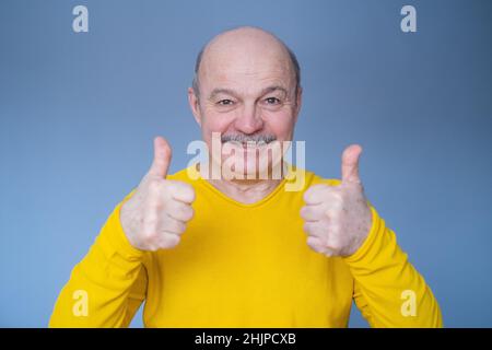 Schöner älterer Mann in gelbem Pullover zeigt Daumen nach oben. Studio auf blauer Wand. Stockfoto
