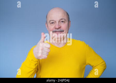 Schöner älterer Mann in gelbem Pullover zeigt Daumen nach oben. Studio auf blauer Wand. Stockfoto