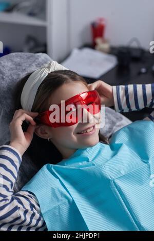 Das kleine Mädchen sitzt in einem Zahnarztstuhl und trägt eine Schutzbrille. Sie dubelt Stockfoto