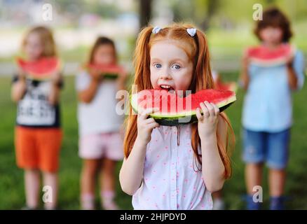 Das rothaarige Mädchen isst gierig saftig reife Wassermelone auf dem Hintergrund ihrer Freunde und parkt an einem sonnigen Sommertag Stockfoto