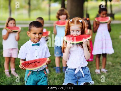 Eine Gruppe von glücklichen Vorschulkindern verschiedener ethnischer Gruppen hält reife Wassermelonen in Scheiben und lächelt im Hintergrund des Parks auf einem Sonnenbad Stockfoto