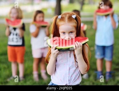 Das rothaarige Mädchen isst gierig saftig reife Wassermelone auf dem Hintergrund ihrer Freunde und parkt an einem sonnigen Sommertag Stockfoto