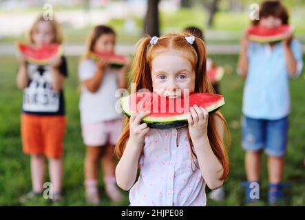 Das rothaarige Mädchen isst gierig saftig reife Wassermelone auf dem Hintergrund ihrer Freunde und parkt an einem sonnigen Sommertag Stockfoto