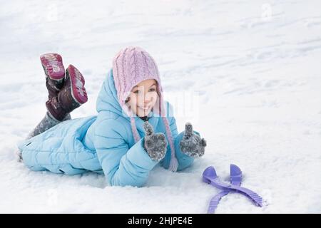 Porträt eines Mädchens im Winter in einem rosa Hut und einer blauen Jacke. Mädchen liegt auf dem Schnee. Mädchen spielt im Schnee Stockfoto