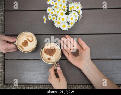 Männliche und weibliche Hände halten Tassen Kaffee, in denen Herzen aus Milch und Zimt hergestellt werden. Liebeskonzept. In der Handfläche liegt Schokolade Süßigkeiten. Moment der Stockfoto