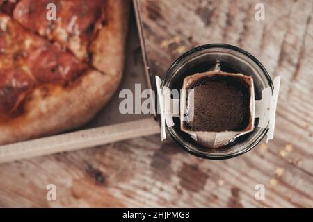 Kaffee in einem Tropfbeutel in einem transparenten Glas auf einem Holzhintergrund zubereiten. Heißer, gemahlener Tropfkaffee, Draufsicht. Äthiopischer Kaffee in einem wiederverwendbaren d gemahlen Stockfoto