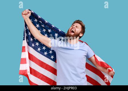 Porträt eines entzückten, zufriedenen bärtigen Mannes, der mit großer amerikanischer Flagge die Ehre hatte, während er den Nationalfeiertag feierte, aufblickte und schrie. Innenaufnahme des Studios isoliert auf blauem Hintergrund. Stockfoto