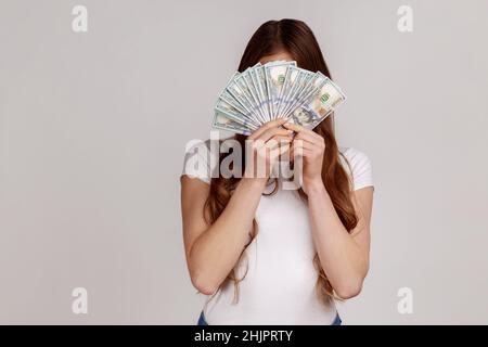 Unbekannte Frau, die sich hinter einem Haufen Dollar-Banknoten versteckt, anonyme Person, die Geld hält, Lottogewinn, großer Gewinn, weißes T-Shirt trägt. Innenaufnahme des Studios isoliert auf grauem Hintergrund. Stockfoto