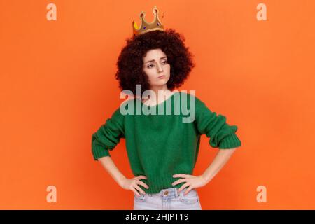 Selbstbewusste Frau mit Afro-Frisur trägt einen grünen Pullover im lässigen Stil und eine Krone, die mit egoistischem Ausdruck steht und die Hüften der Hand hält. Innenaufnahme des Studios isoliert auf orangefarbenem Hintergrund. Stockfoto