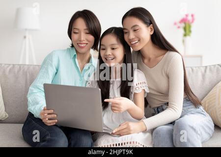 Lächelndes asiatisches Mädchen mit ihrer Mutter und Oma beim Ansehen von Videos und beim Suchen im Internet auf dem Laptop im Wohnzimmer Stockfoto