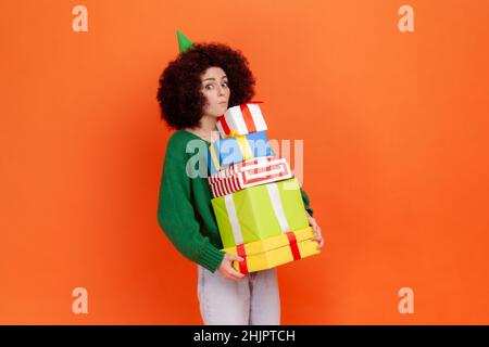 Erstaunt Frau mit Afro-Frisur trägt grünen lässigen Stil Pullover stehen mit Stapel von Geschenken in den Händen, erstaunt, viele Geschenke zu haben. Innenaufnahme des Studios isoliert auf orangefarbenem Hintergrund. Stockfoto