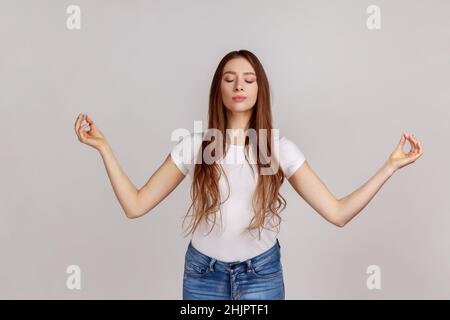 Porträt einer ruhigen, schönen Frau, die die Hände in Mudra-Geste hochhält, die Augen während der Meditation geschlossen hält, Yoga praktiziert, weißes T-Shirt trägt. Innenaufnahme des Studios isoliert auf grauem Hintergrund. Stockfoto