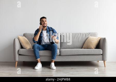 Fröhlicher arabischer Mann, der auf dem Handy spricht und auf der Couch sitzt Stockfoto