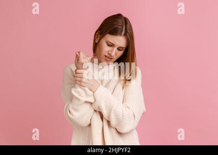 Porträt einer blonden Frau, die mit Schmerzen steht, schmerzende Handgelenke massiert, Handverletzungen oder Verstauchungen leidet und weißen Pullover trägt. Innenaufnahme des Studios isoliert auf rosa Hintergrund. Stockfoto