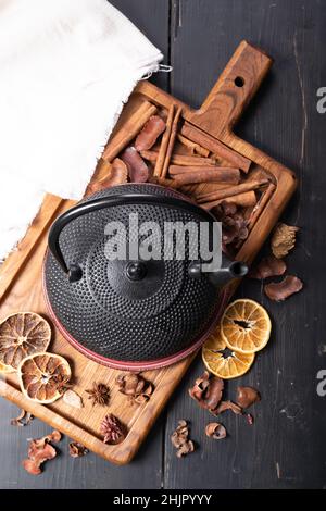 Schwarze Teekannen aus Gusseisen. Zimtstangen, Sternanisgesteine, trockene Orangenscheiben und getrockneter Ingwer auf einem Schneidebrett. Draufsicht. Vertikale Aufnahme Stockfoto