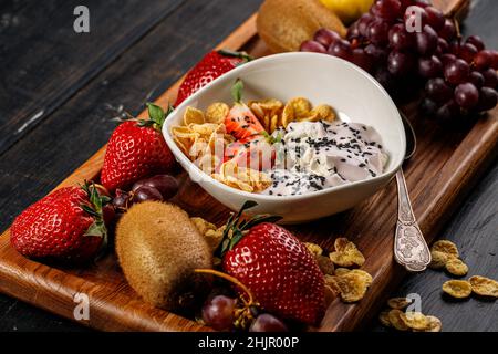 Maisflocken mit Joghurt und Chiasamen. Verschiedene saftige reife Früchte. Erdbeere, Kiwi, Apfel, Birne, Banane, Trauben. Köstliche tropische Früchte auf einem Holz Stockfoto