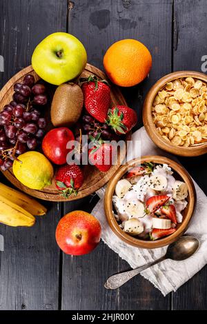 Maisflocken mit Joghurt und Chiasamen. Verschiedene saftige reife Früchte. Erdbeere, Kiwi, Apfel, Birne, Banane, Trauben. Köstliche tropische Früchte auf einem Holz Stockfoto