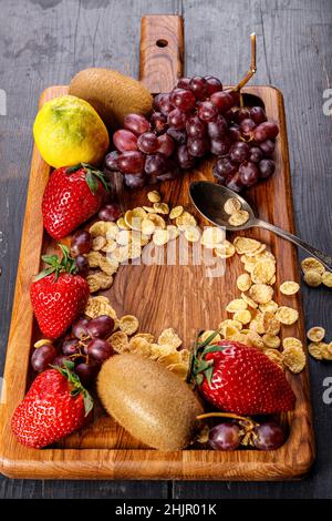 Cornflakes. Verschiedene saftige reife Früchte. Erdbeere, Kiwi, Trauben. Köstliche tropische Früchte auf einem hölzernen Hintergrund. Gesundes Frühstück. Vertikale Aufnahme. Stockfoto