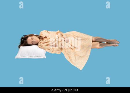 Schlafende Schönheit schwebt in der Luft. Entspanntes Mädchen in gelbem Kleid liegt bequem auf Kissen schweben, die Augen geschlossen halten, beobachten Träume. Volle Länge Studio Schuss isoliert auf blauem Hintergrund, innen Stockfoto