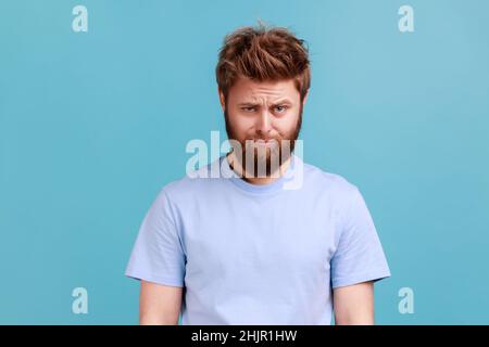Porträt eines bärtig verärgerten jungen Mannes, der mit unzufriedenem Traurigkeit ins Gesicht steht und die Kamera anschaut, Trauer ausdrückt, schlechte Laune hat. Innenaufnahme des Studios isoliert auf blauem Hintergrund. Stockfoto