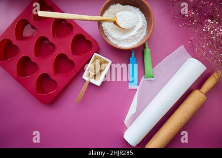 Backvorbereitung: Herzförmige Silikonform, Nudelholz, Süßwarenspritze, Glasur, Mehl, Zucker auf rosa Hintergrund. Valentinstag-Konzept. Hochwertige Fotos Stockfoto