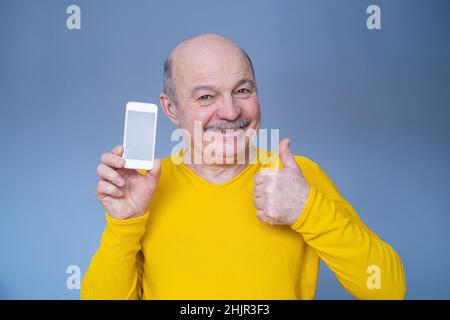 Schöner älterer Mann in gelbem Pullover zeigt Daumen nach oben und Telefonbildschirm mit Copyspace. Studio auf blauer Wand. Stockfoto