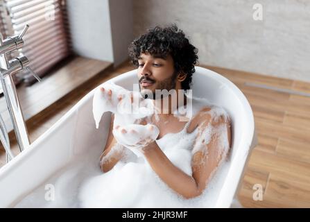 Hübscher indischer Mann bläst Seifenblasen, während er sich in der Badewanne mit Schaumstoff und freiem Platz entspannt Stockfoto