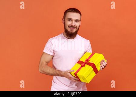 Porträt eines glücklichen, positiven bärtigen Mannes, der in einer gelben Geschenkbox steht, mit einem toothy Lächeln auf die Kamera blickt und ein rosa T-Shirt trägt. Innenaufnahme des Studios isoliert auf orangefarbenem Hintergrund. Stockfoto