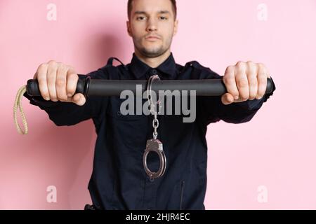 Ein gutaussehender Polizist in schwarzer Uniform mit Keule und Handschellen isoliert auf rosa Hintergrund. Stockfoto