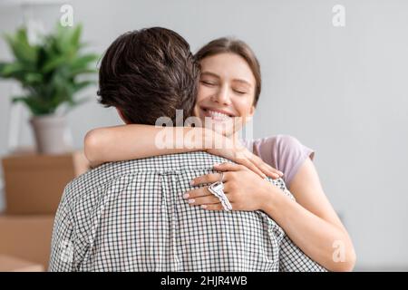 Lächelnd, fröhlich, hübsche junge europäische Frau umarmt ihren Mann und zeigt den Schlüssel aus der neuen eigenen Wohnung im Zimmer Stockfoto