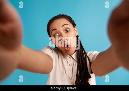 Erstaunt Frau mit schwarzen Dreadlocks hat Gesichtsausdruck überrascht, streckt Arm für Selfie, drückt aufrichtige Emotionen, Standpunkt Foto. Innenaufnahme des Studios isoliert auf blauem Hintergrund. Stockfoto