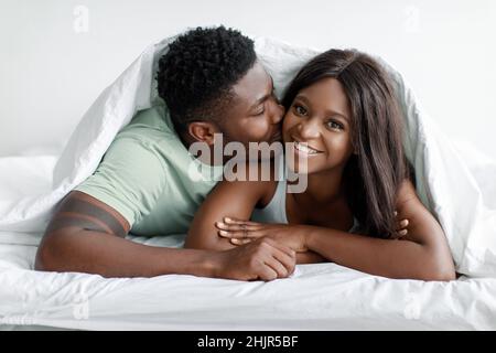 Lächelnder junger afroamerikanischer Ehemann küsst seine Frau auf der Wange unter Bettdecken Stockfoto