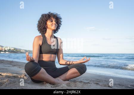 Die schwarze Frau lächelt entspannt, während sie Yoga am Meer praktiziert Stockfoto