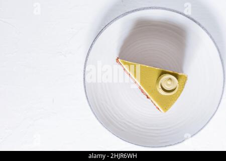 Pistazien-Schokoladenkuchen mit vier Schichten und grüner Pistazien-Verglasung auf einem weißen Teller auf weißem Hintergrund serviert mit Kaffee in Unschärfe Stockfoto