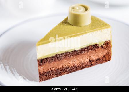 Pistazien-Schokoladenkuchen mit vier Schichten und grüner Pistazien-Verglasung auf einem weißen Teller auf weißem Hintergrund serviert mit Kaffee in Unschärfe Stockfoto