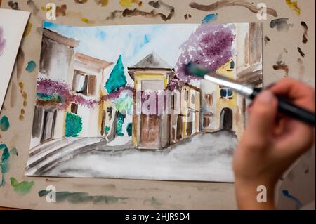 Nahaufnahme des lebendigen Aquarellmalprozesses. Pinsel in der Hand über Malerei. Bunte mittelalterliche Stadtstraße auf Papptafel. Stockfoto