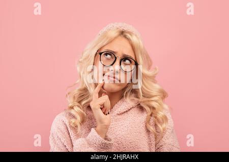 Porträt einer nachdenklichen blonden Dame, die das Kinn berührt und zur Seite schaut, posiert auf pinkem Studiohintergrund Stockfoto