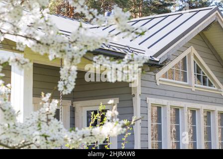 Schönes Holzhaus mit großen Fenstern, einem Schaufenster und einer Terrasse im Hintergrund blühender Frühlingsbäume Stockfoto