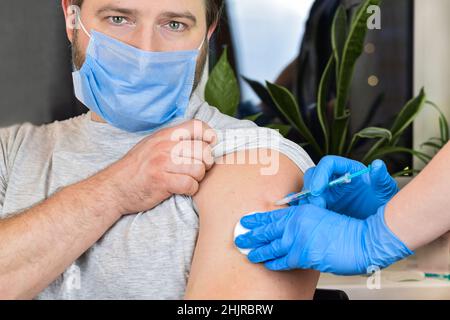 Hände eines Arztes, der Coronavirus-Covid-19-Impfstoff in eine Impfspritze in den Armmuskel des kaukasischen Mannes zur Covid-Immunisierung injiziert. Stockfoto