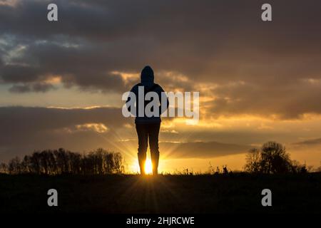 Rückansicht Silhouette einer isolierten Frau in Wintermantel & Gummistiefel auf einem Spaziergang auf dem Land, Blick auf den Sonnenuntergang Himmel, beobachten den Sonnenuntergang mit Sonneneinstrahlung. Stockfoto