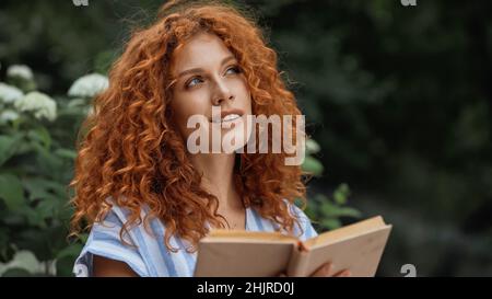 Verträumte Rothaarige Frau lächelt, während sie Buch im Freien hält Stockfoto