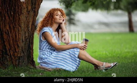 Verträumte Rothaarige Frau in blauem Kleid lächelt, während sie unter dem Baumstamm sitzt und Kaffee hält Stockfoto
