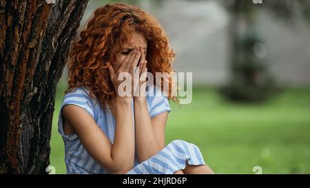 Traurige junge Frau mit roten Haaren, die das Gesicht bedeckt, während sie im Park weinte Stockfoto
