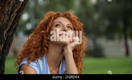 Verträumte Frau mit lockigen Haaren, die im Park aufschaut und lächelt Stockfoto