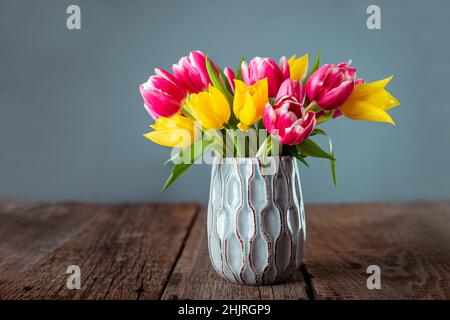 Frischer Frühlingsbouquet aus gelben und rosa Tulpen in blauer Vase auf rustikalem Holztisch mit grauem Hintergrund. Festliche Blumen für Mütter oder Frauen Stockfoto
