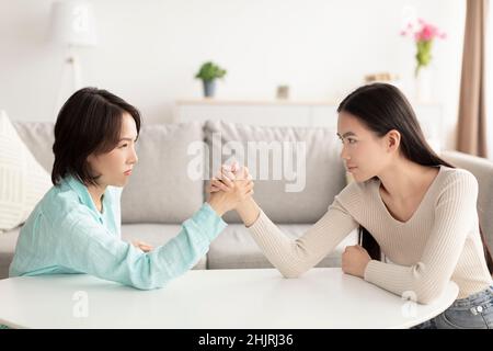 Reife asiatische Mutter und Erwachsene Tochter Arm Wrestling, einander in die Augen schauen, kämpfen zu Hause Stockfoto